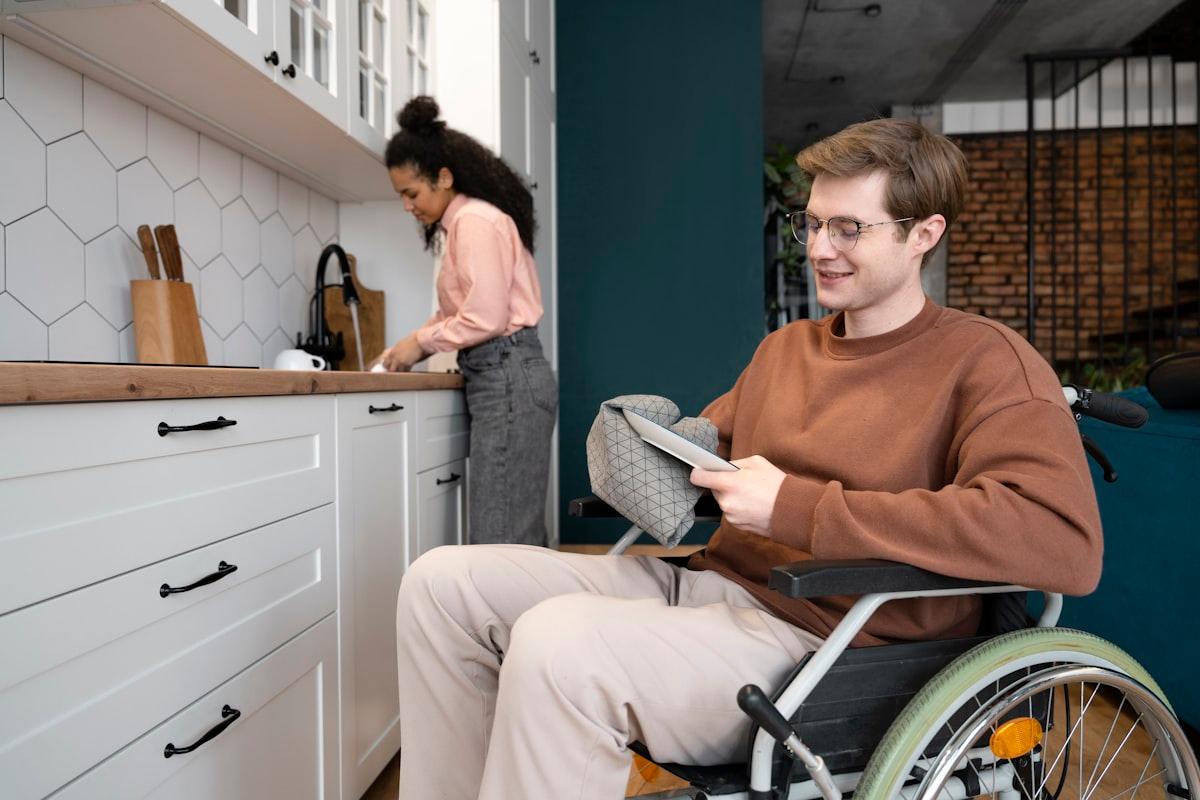 A man in a wheelchair at home in his kitchen while a support worker prepares food